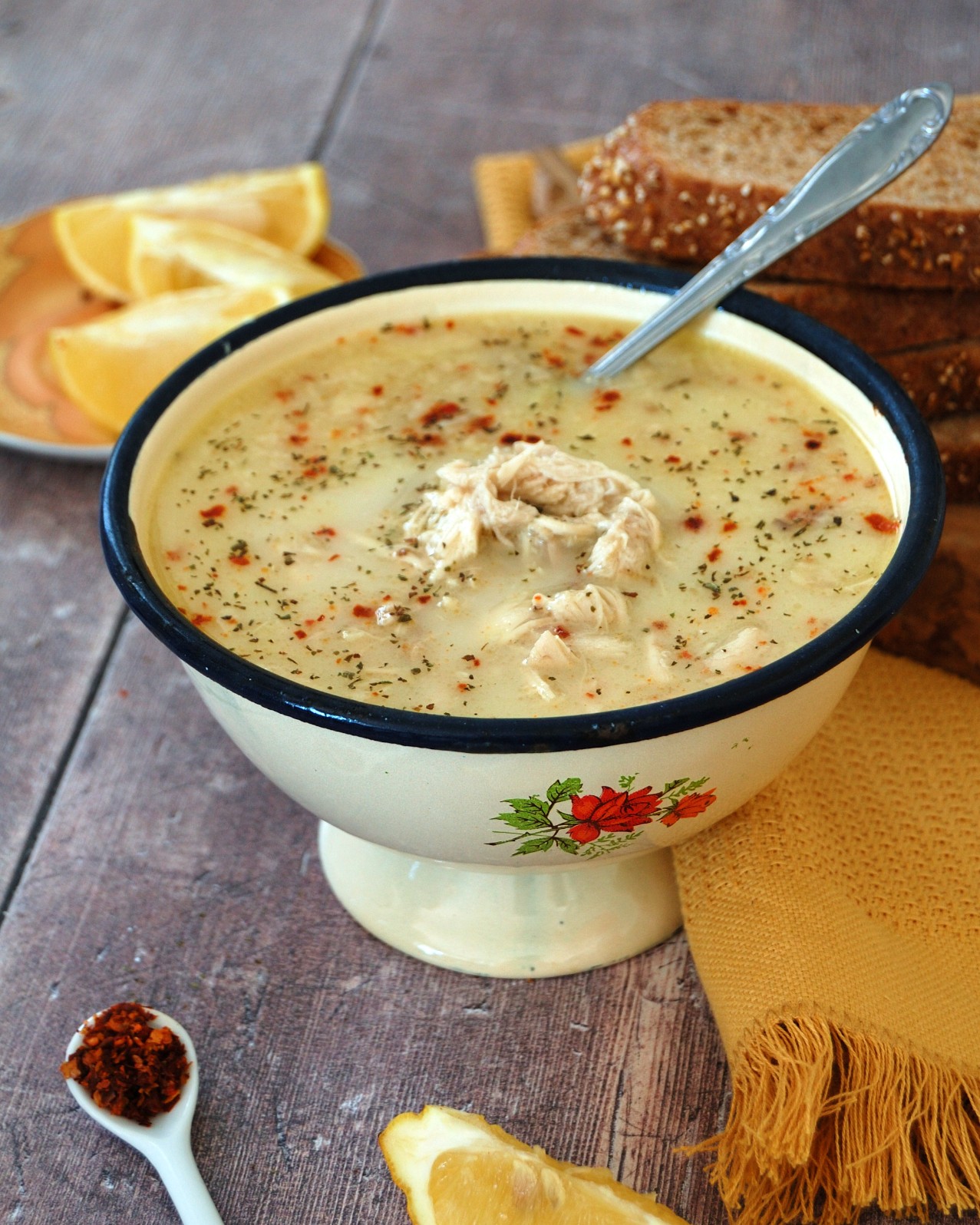 Vintage enamel bowl with sliky cream coloured chicken soup recipe. Lemon wedges and sliced brown bread to rear. Shredded chicken and pepper flakes can be seen on top of the Turkish soup