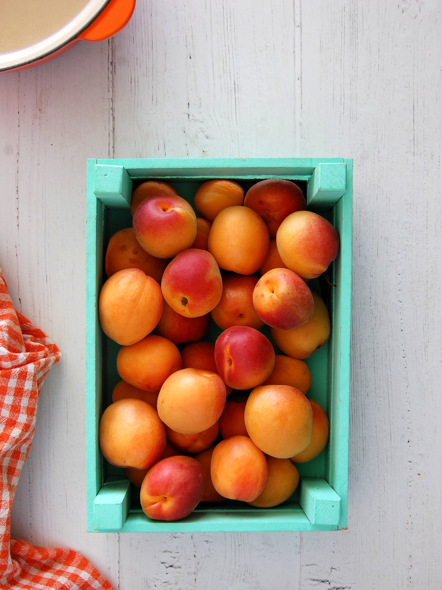 Tourquoise green crate holding fresh apricots. Top left cast iron orange saucepan on a wooden background