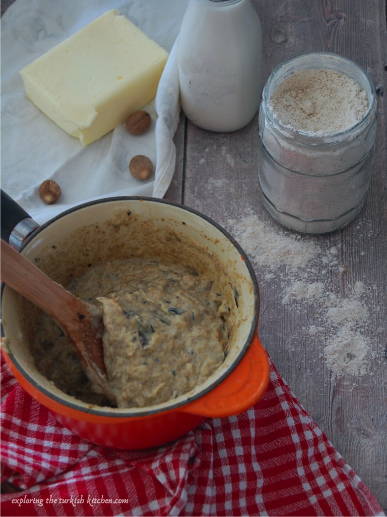 A small le cruset saucepan with aubergine based sauce sits on a red checkered tea towel. Behind it wait a open jar of wholewheeat flour, a dairy bottle of milk and a block of butter on a wap with nutmeg seeds. 