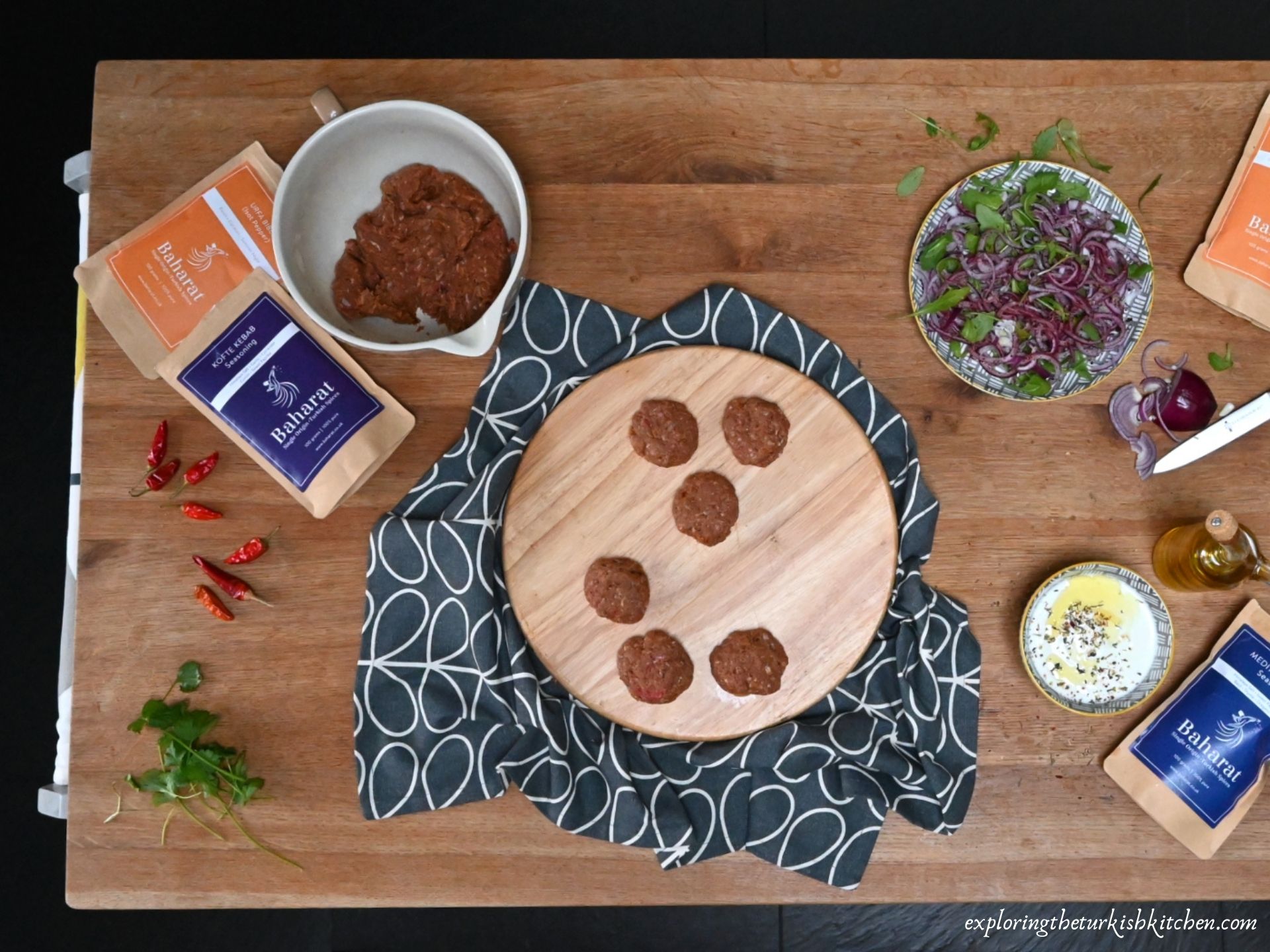 A large butchers block. Scene shows making Turkish meatballs. Packets of Baharat brand spices mixes, olive oil, chillies and yoghurt surround some made meatballs and unmade mixture