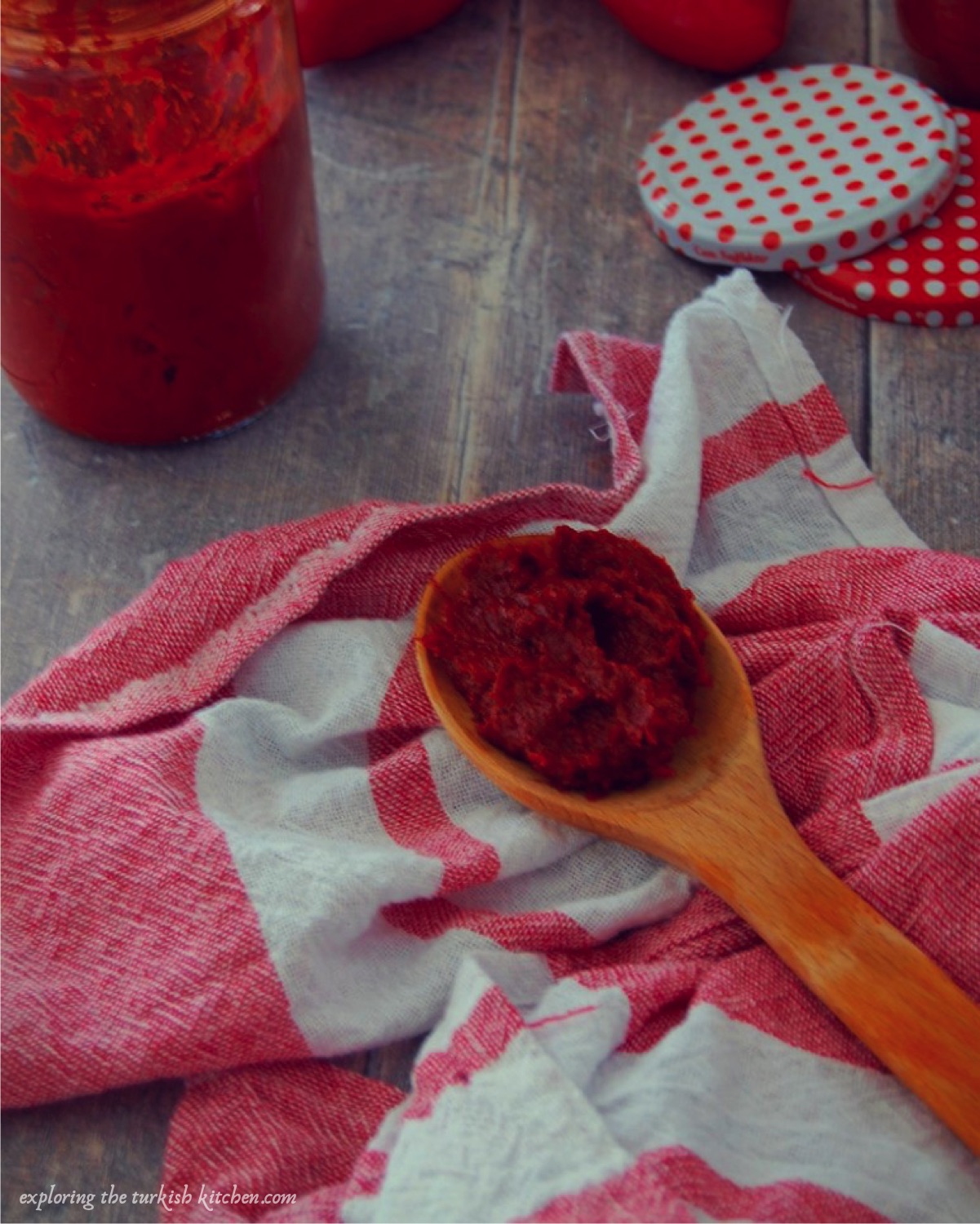 Large wooden spoon with Turkish sundried red pepper paste lays on a red and white teatowel. A half filled jar and red polka dot lid set the scene.