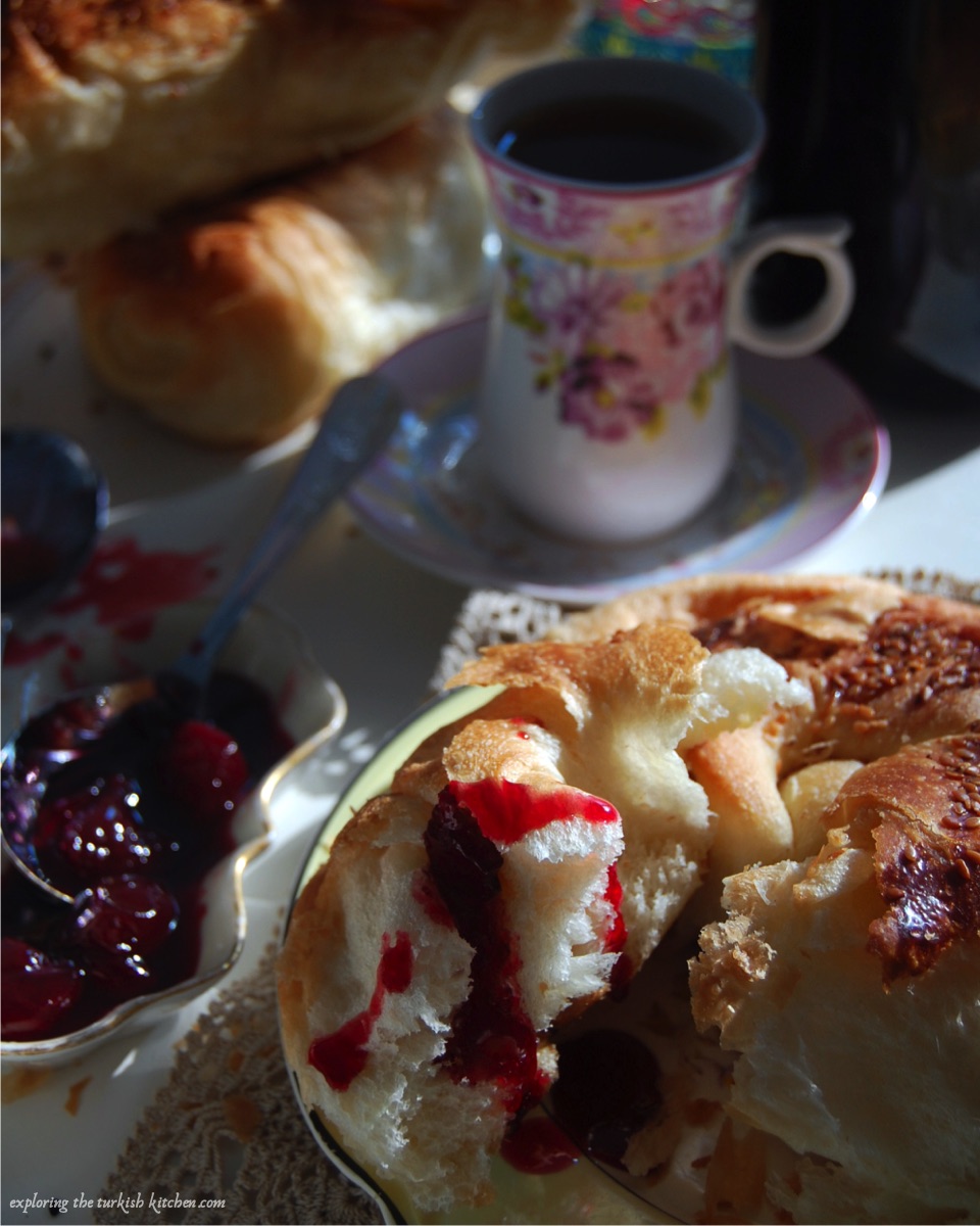 Turkish soft bread roll, açma drizzled with sour cherry jam. Smaşş bowl of jam and spoon and floral Turkish tea glass for a Turkish quick breakfast