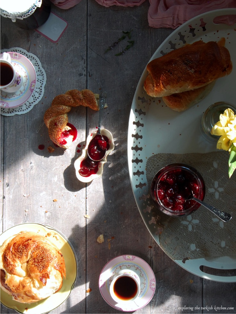 Breakfast scene. Wooden table scattered with crumbs and cream decorative tray. Bread rolls and simits scatted with spoons of sour cherry jam. Pink floral Turkish tea glasses. Sun casts accross the table onto the jam and bread  