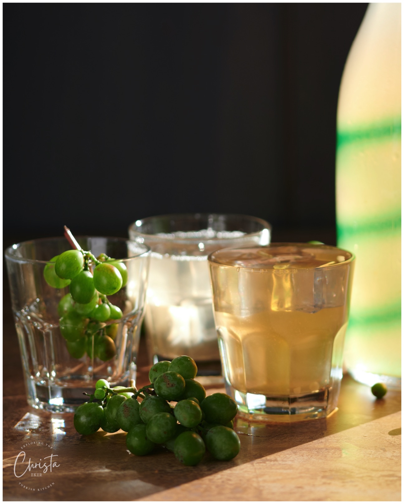 Picture shows a bottle of homemade koruk drink Turkish Koruk şerbeti and glasses of juice and one filled with unripe grapes.