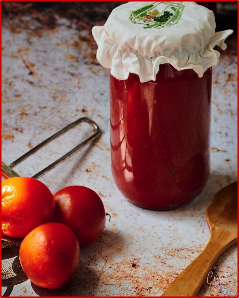 Jar of Turkish tomato paste 'Salça' next to a small colander of plum tomatoes. The sun rays shin on the tomatos and onto the jar of Domates salça