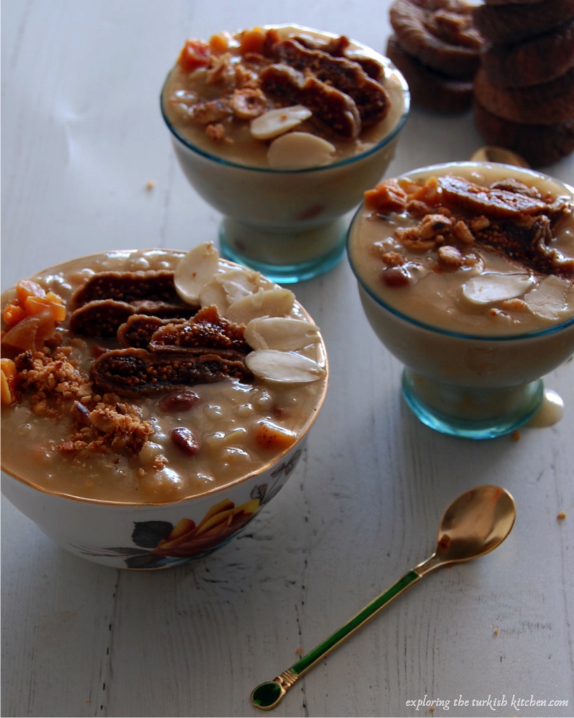Small bowls of ancient grain pudding topped with sliced dried figs, crushed and blanched nuts and a sprinkling of cinnamon. Golden and green jewel spoons aside. A stack of dried figs in the background.