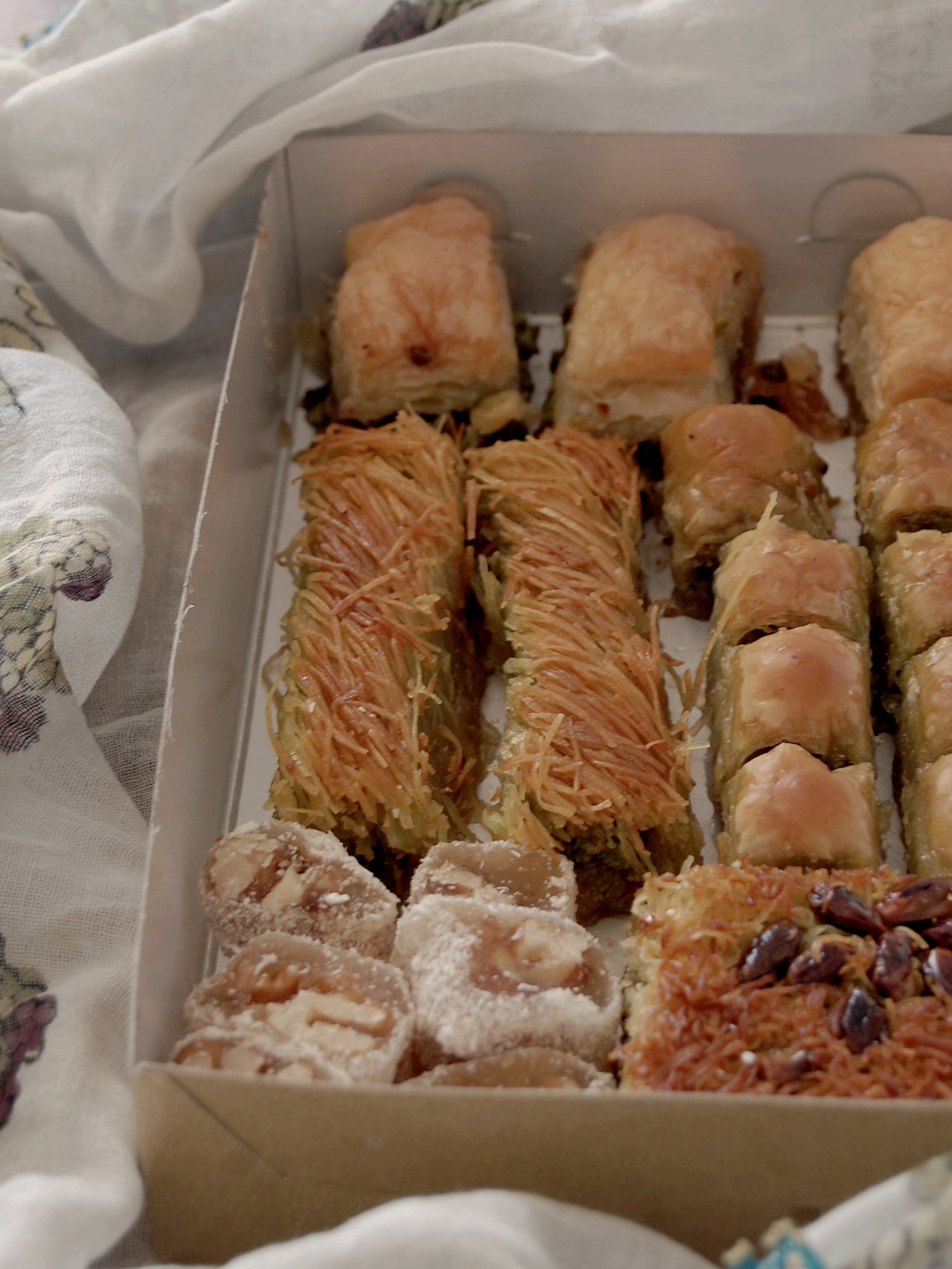 Turkish desserts in a box surrounded by light blue headscarf. Box holds large peices of baklava and rear. Burma kadayıf strands bankalava, mini baklava, and turkish delight with walnuts at the front.