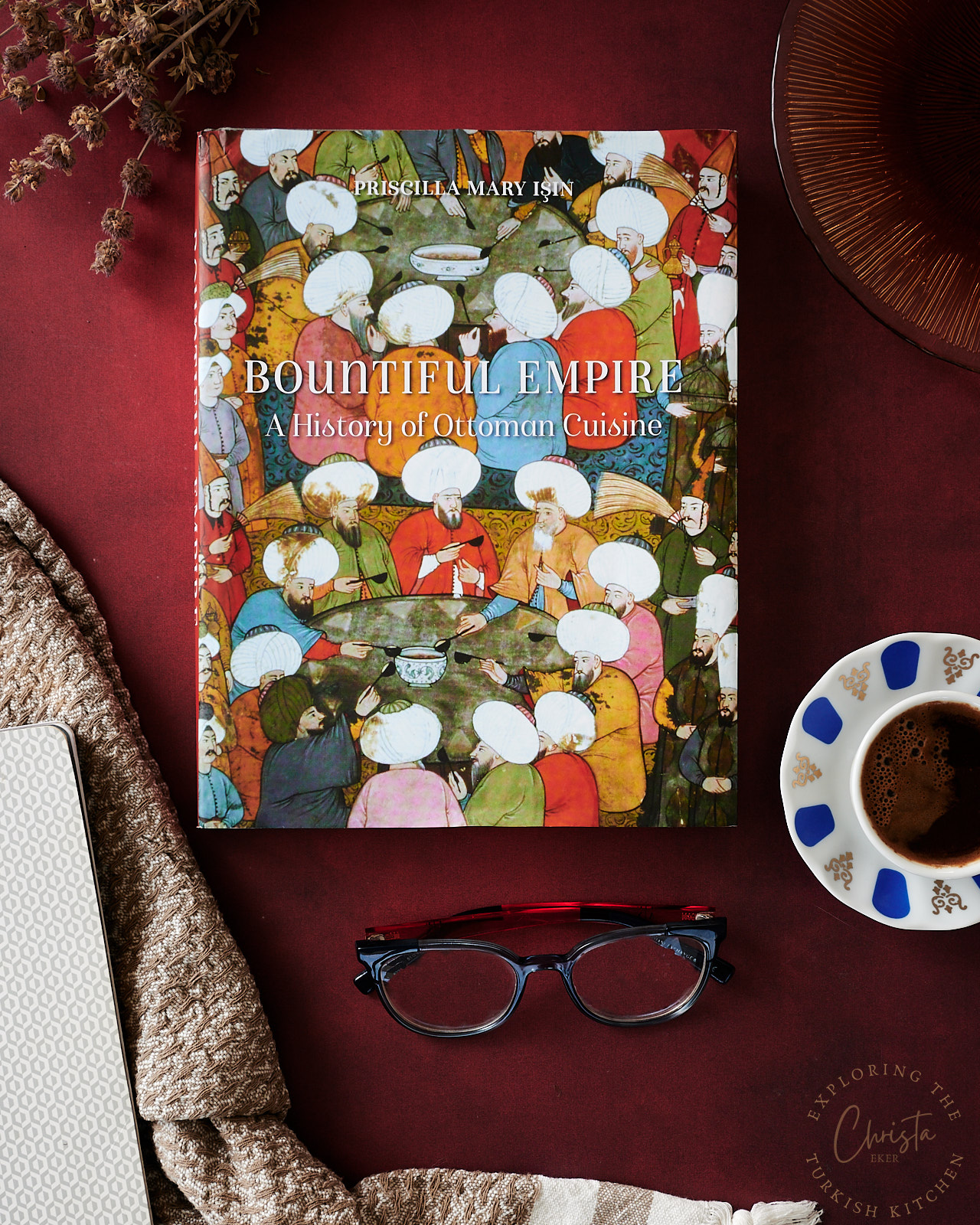 Moody book scene: There is a deep red background. In the Centre of frame is Mary Işın's Book surrounding it clockwise tried thyme, a handmade gold glass plate, Turkish coffee, a pair of glasses and a brown and white scarf topped with a gold and white note book.