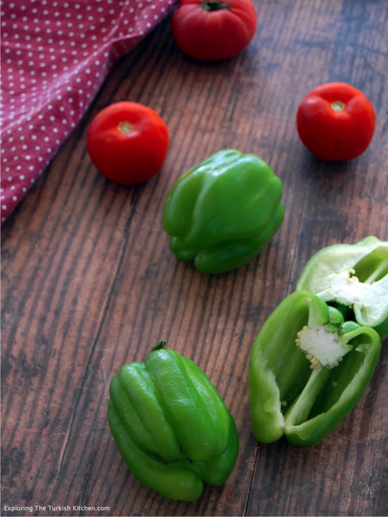 Two green bell peppers, one cut in half and 3 small tomatoes with a red checkered tea towel on a wooden board.