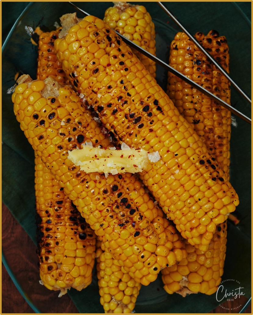 Picture of Turkish corn on the cobs grilled using a kozmatik hob plate. A tong sits on the cobs that have scrorched kernals and are a piece of butter and pepper flakes on them
