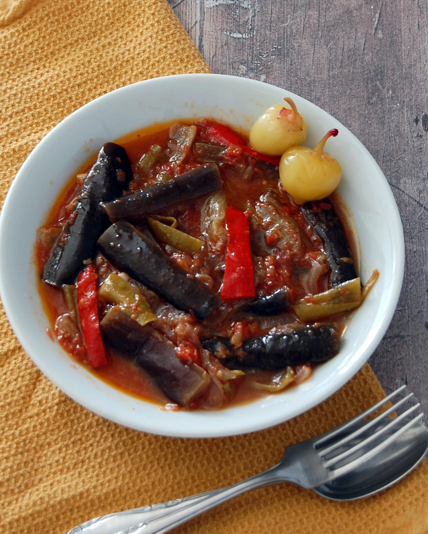 White bowl with wedges of aubergine and slices of red and green pepper in a tomato sauce. Bowl placed on a mustard cloth and wooden background. Decorate cutlery below. 