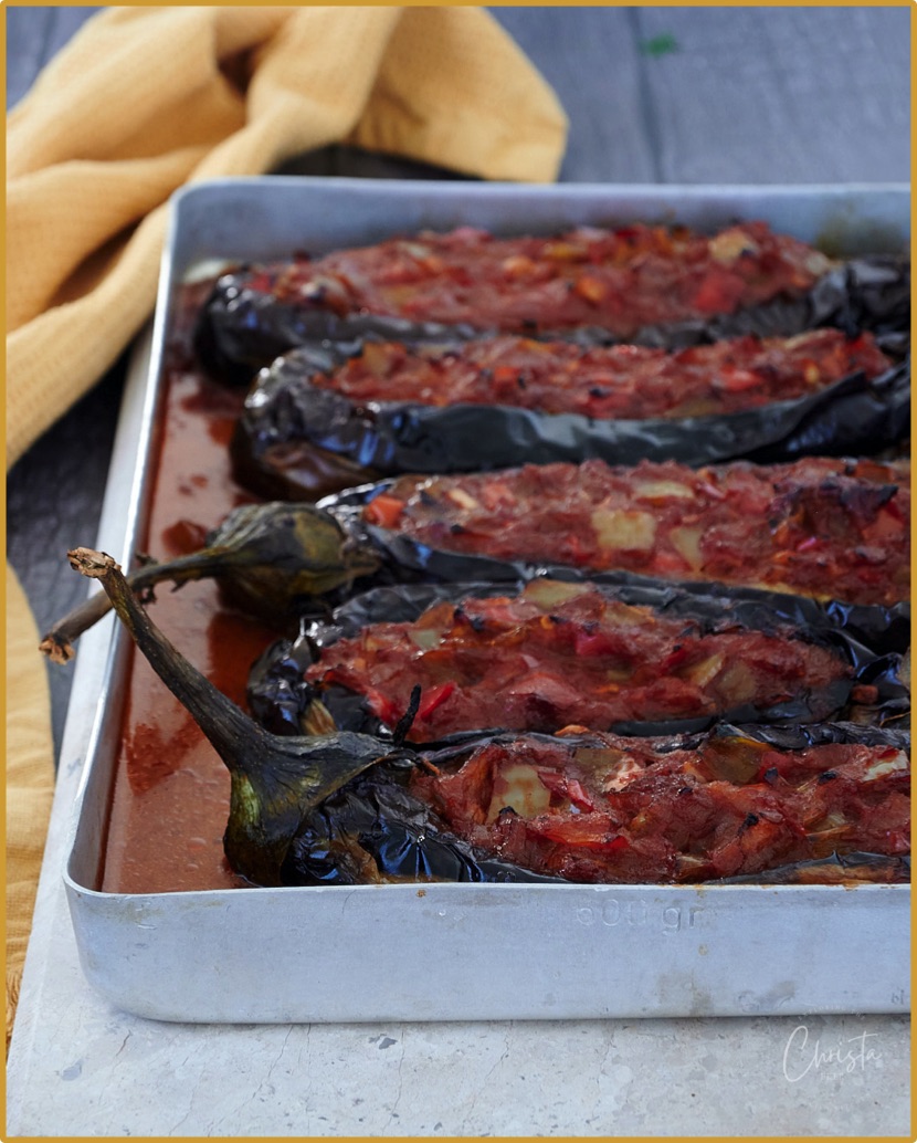 Rectangle tray on a marble slab. Cooked stuffed aubergines are lined up in the tray with a tomato sauce. Mustard tea-towel sits on the left. 